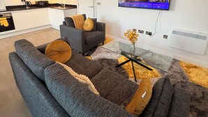 A modern living room with a grey sectional sofa, a glass coffee table, and a flat-screen TV mounted on the wall. - Grace House (St Albans)