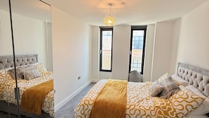A bedroom with a large mirror, a bed with patterned bedding, a hanging light fixture, and a window with a view. - Grace House (St Albans)