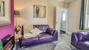 A living room with a purple leather sofa, a small side table with a vase of flowers, a painting on the wall, and a door leading to another room. - Corner House, close to Lincoln City Centre (Lincoln)