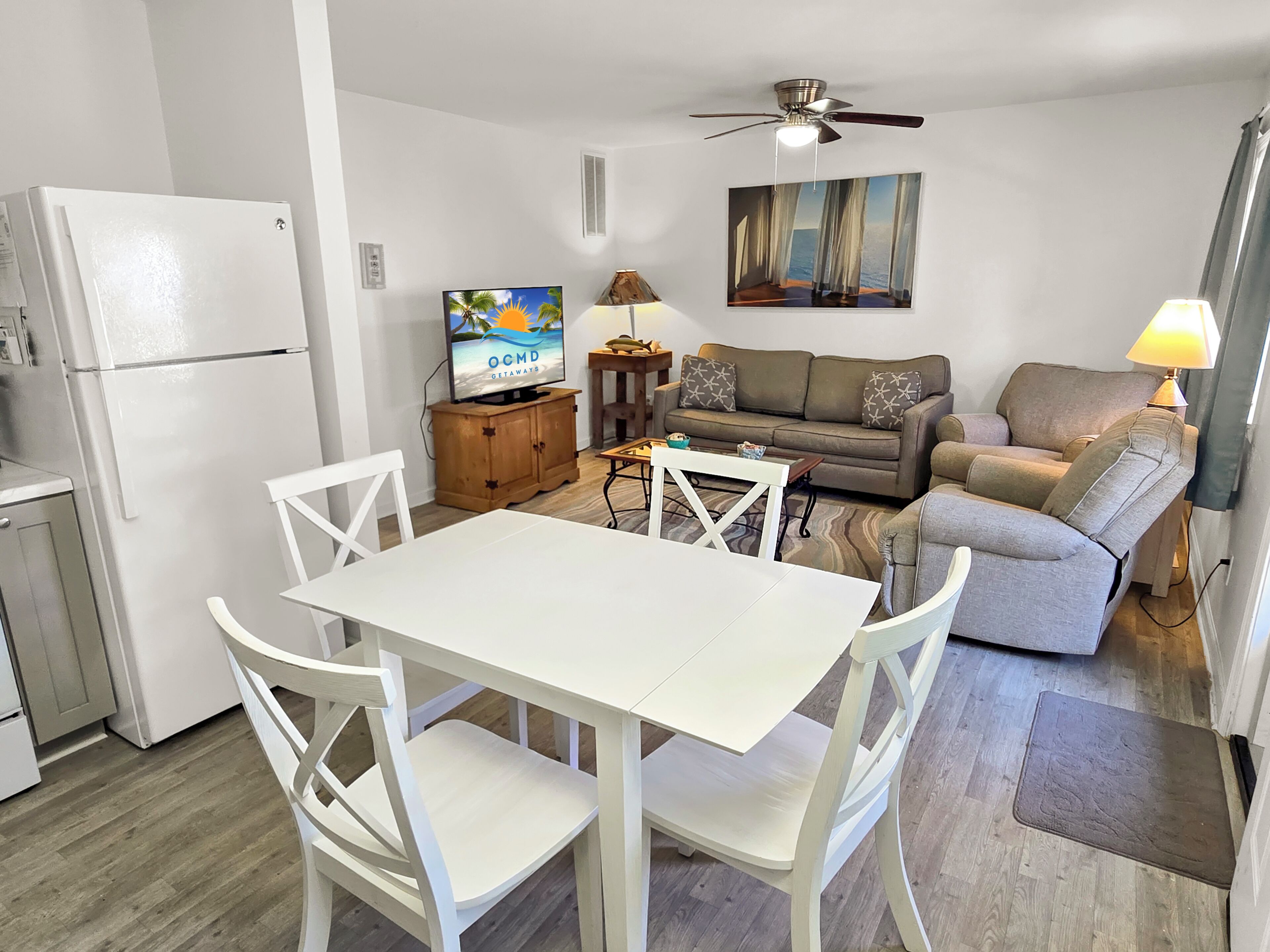 A compact living space with a kitchenette, a dining area with a white table and chairs, a sofa, a TV, and a ceiling fan.
