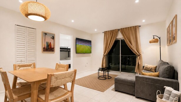 Een moderne woonkamer met een eethoek, een bank, een televisie en een groot raam met gordijnen.