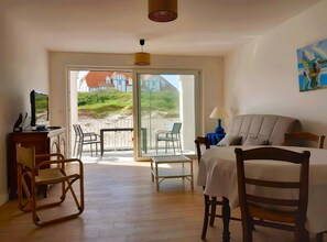 Una habitación con una cama, un escritorio con televisión, una silla y una mesa. Se ve una playa y casas a través de una puerta de vidrio.
