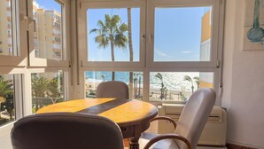 A room with a dining table, chairs, a view of the beach, and a window with a view of palm trees and buildings. - Apartment 'Beach Front Home A125' with Sea View, Wi-Fi and Air Conditioning (Torrox Costa)