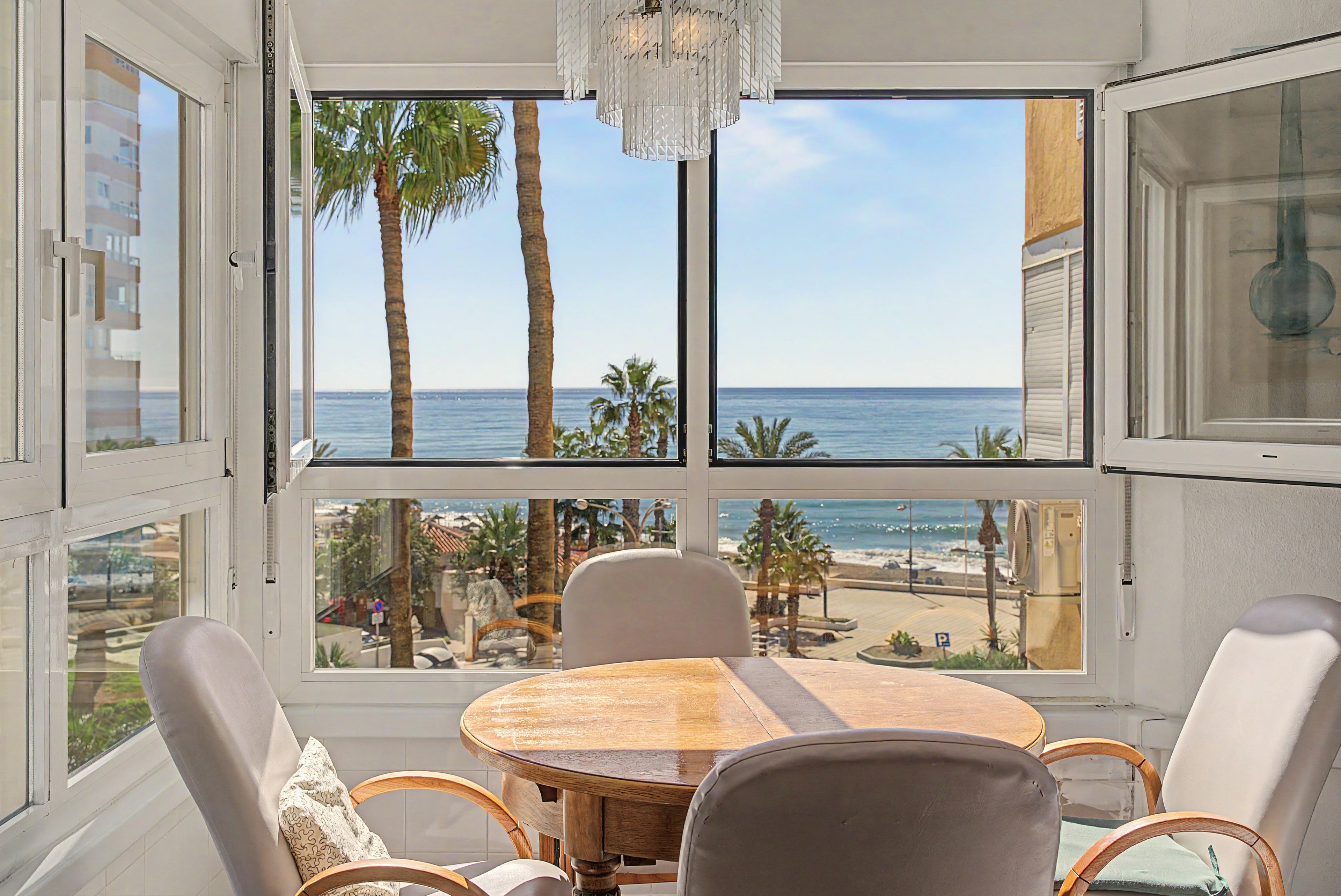 Ein Zimmer mit einem großen Fenster mit Blick auf einen Strand, einem runden Holztisch und Korbstühlen.
