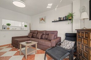 A living room with a brown sectional sofa, a small table, a chair, a bookshelf, and a wall-mounted clock. - Apartment 'Beach Front Home A125' with Sea View, Wi-Fi and Air Conditioning (Torrox Costa)
