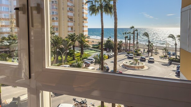 A view from a window overlooking a beach, palm trees, and a parking area with cars and a motorcycle. - Apartment 'Beach Front Home A125' with Sea View, Wi-Fi and Air Conditioning (Torrox Costa)