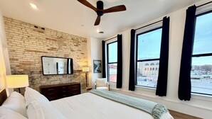 Un dormitorio con pared de ladrillo, ventanas amplias, ventilador de techo, cama con ropa blanca, cómoda y lámpara.