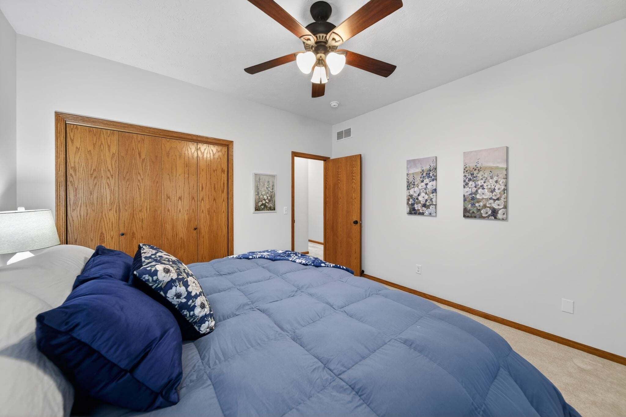 A bedroom with a bed, wooden wardrobe, ceiling fan, and wall-mounted artwork.