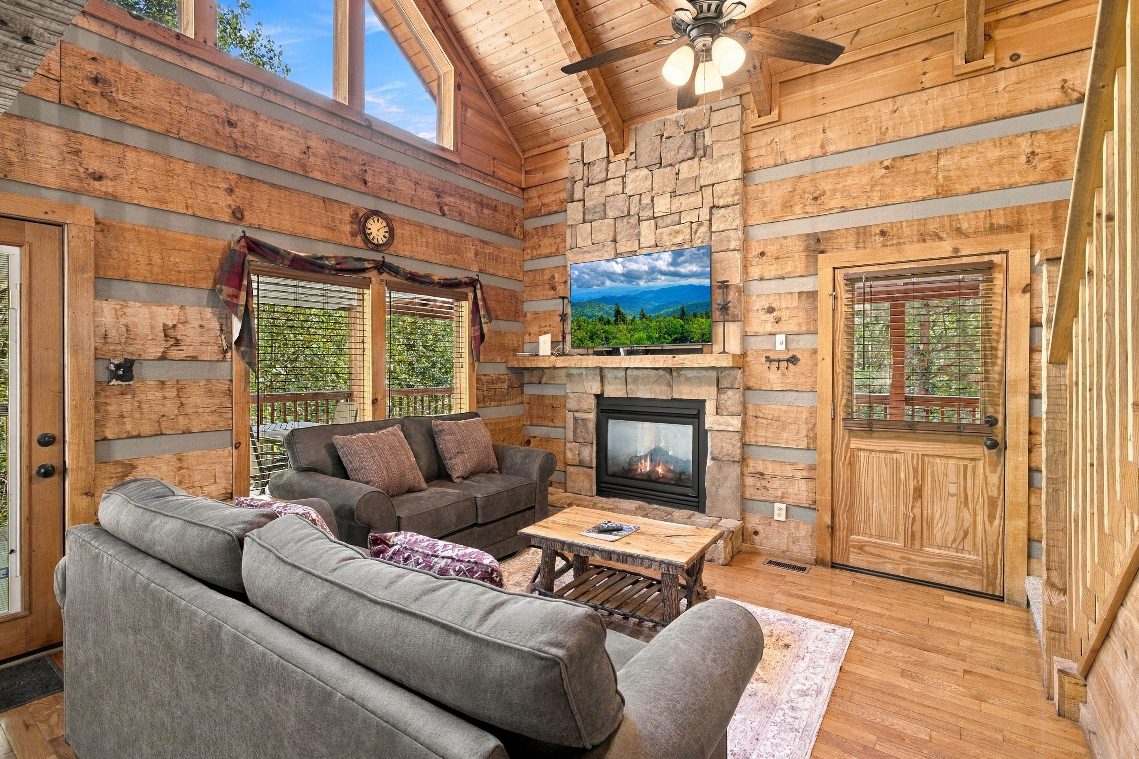 A log cabin interior with a stone fireplace, a sofa, a television, and a ceiling fan.