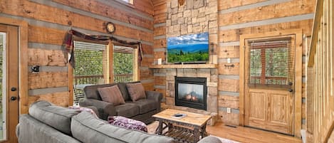 A log cabin interior with a stone fireplace, a sofa, a television, and a ceiling fan.