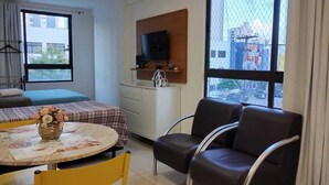 Habitación de hotel con cama, escritorio con televisor, mesa de comedor con sillas y ventanas amplias que ofrecen vistas de la ciudad.