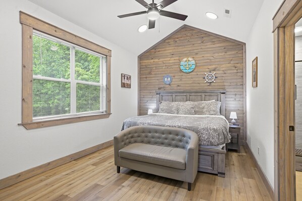 A bedroom with a large window, a ceiling fan, a bed with a headboard, a bedside table, and a chair. - Sanctuary Retreat 3 Bedroom Home (Sevierville)