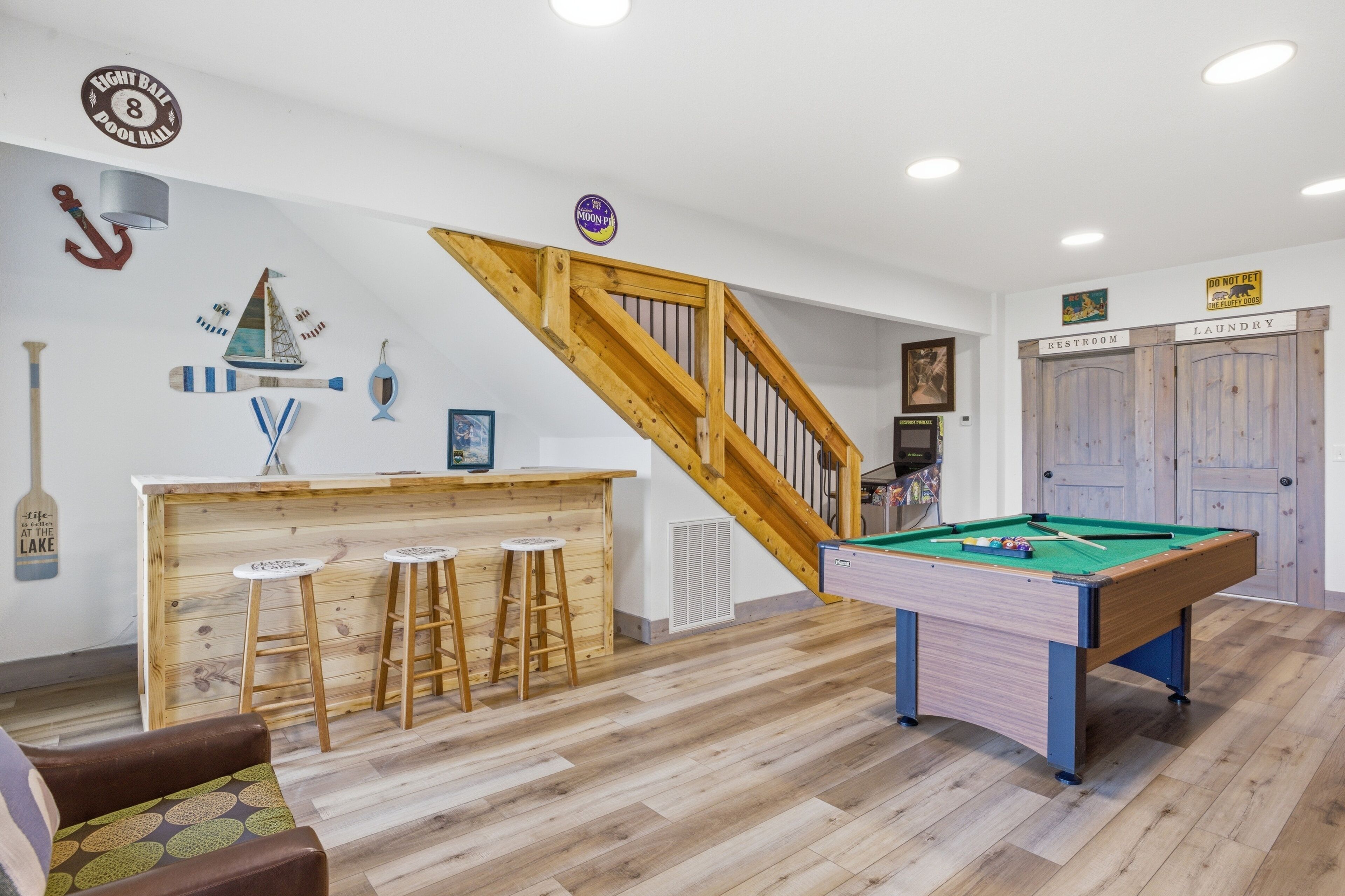 A pool table, bar area, and wooden staircase in a room with white walls and wooden flooring.