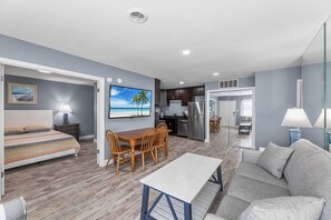 A modern living room with a sofa, a dining table with chairs, a kitchen area with a refrigerator, and a bedroom with a bed and nightstand. - Club Ocean Villas 63 (Ocean City)