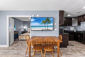 A dining area with a wooden table and chairs, a painting of a beach scene, and a kitchen with dark cabinets. - Club Ocean Villas 63 (Ocean City)