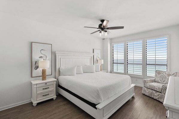 A bedroom with a white bed, a nightstand, a chair, a ceiling fan, and a window with blinds. - Unit 0703E a beautiful 2 bedroom and 2 bath unit (Orange Beach)