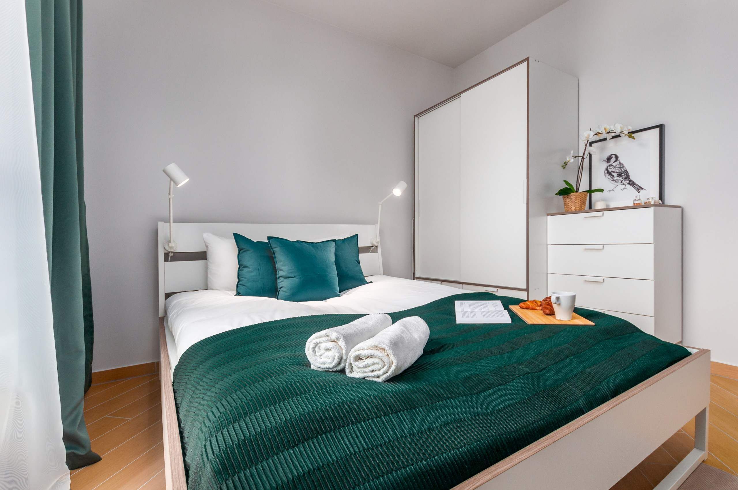 En pænt redt seng med turkise puder, et grønt quilt og sammenrullede håndklæder. Der er et natbord med en lampe, et indrammet billede og en lille plante.