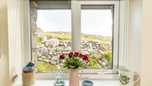 Una ventana con vista a un muro de piedra y follaje, un jarrón con flores rojas, un frasco pequeño, una botella y una jarra decorativa.
