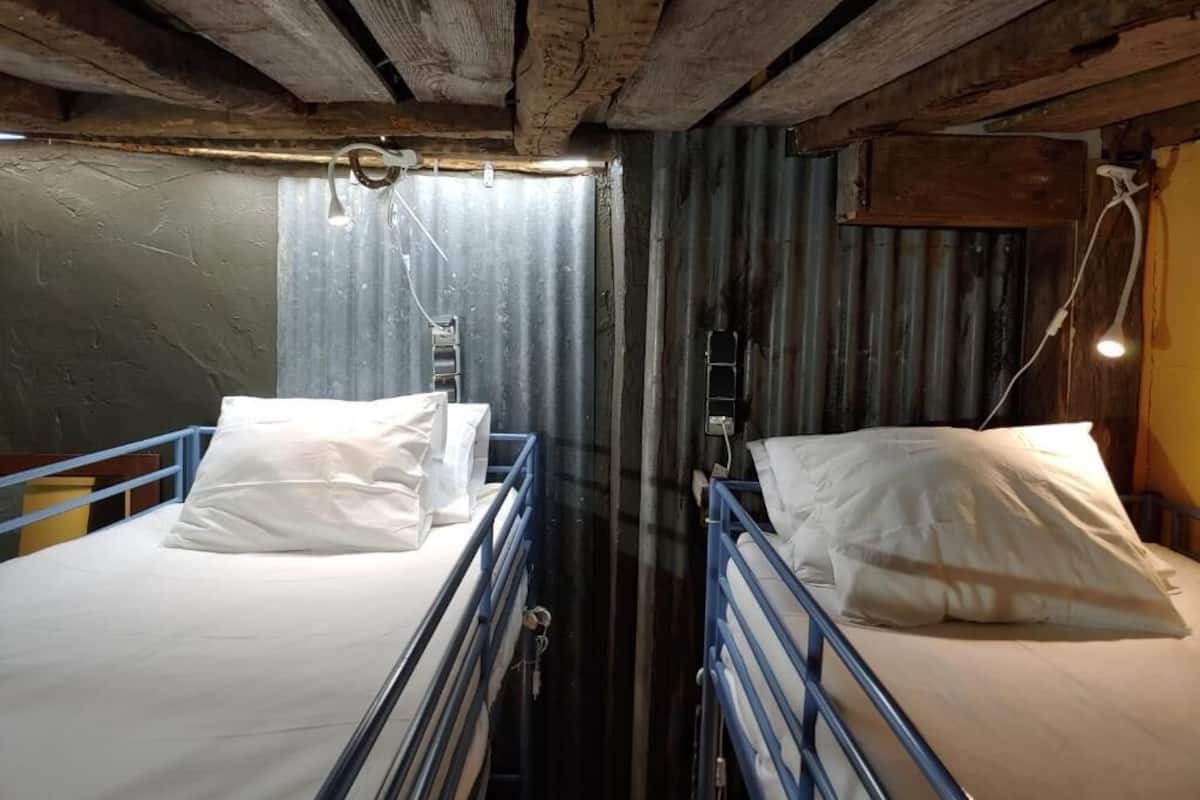 Una habitación estrecha con dos literas, barandillas de metal, techo de madera y pared de metal corrugado.