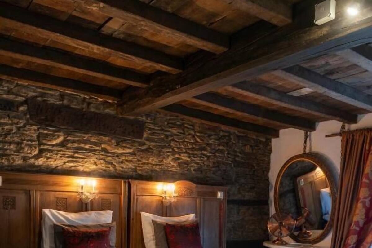 Un dormitorio con pared de piedra, vigas de madera, cama con ropa blanca y un espejo redondo.