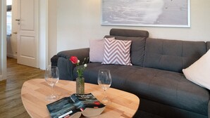 Un salon avec un canapé gris, une table basse en bois sur laquelle reposent deux verres à vin et un livre, un vase rempli de fleurs et une décoration murale sur le thème de la plage.