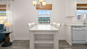 A dining area with a white table and chairs, a chandelier, a window with blinds, and a kitchen area with a dishwasher. - Great Location for Family fun at Jolly Roger and Boardwalk! (Ocean City)