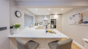 A modern kitchen with a central island, built-in appliances, and a dining area with two chairs. - Beautifully Converted Workshop Close to the Beach | Gweithdy-Bronafon (Borth y Gest)