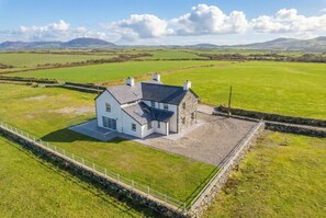 Une maison avec une extension en pierre, entourée d’un mur de pierre et d’une clôture blanche, située dans un champ verdoyant, avec des collines qui se dessinent à l’horizon.