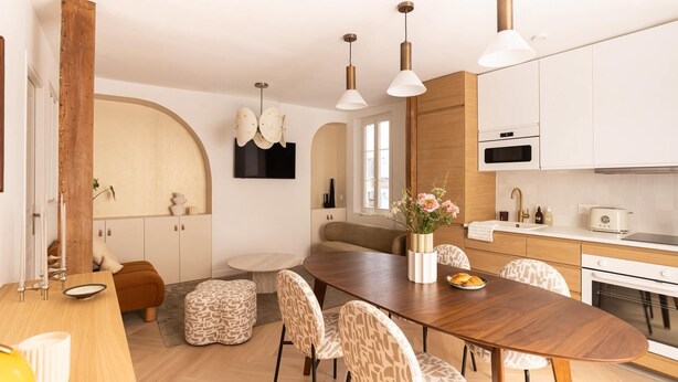 A modern kitchen with white cabinets, a built-in oven, and a dining area with a wooden table and patterned chairs. - Appartement 2 chambre la fayette gare du nord - 4p (Paris)