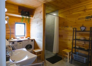 Une salle de bain rustique, aménagée dans une cabane en bois, comprenant une machine à laver, un lavabo, une douche et une étagÚre avec des articles de toilette.