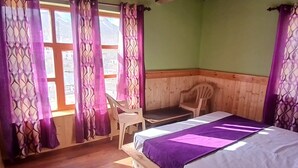 Ein Zimmer mit einem Holzbett, einem Sessel, einem Fenster mit violetten Vorhängen und einer Holzbalkendecke.