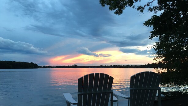 Two Adirondack chairs facing a lake with a sunset.