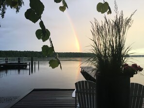 A dock with a chair, a potted plant, and a view of a lake with a rainbow.