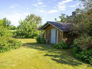 A wooden shed with a door and a small porch, surrounded by lush greenery and a grassy lawn. - 6 Person Holiday Home in Samso (Samsø)