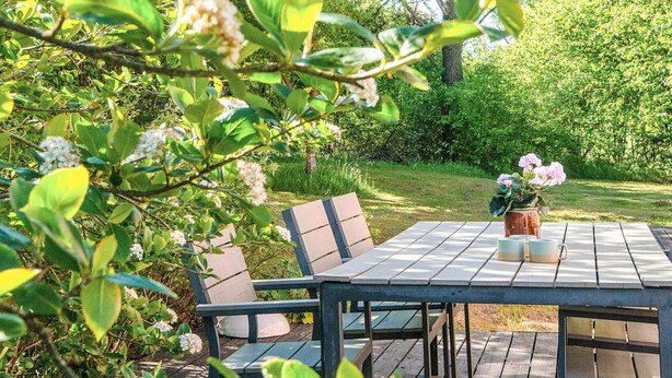 Eine Holzterrasse im Freien mit Tisch und Stühlen, umgeben von Grünpflanzen und Blumen.