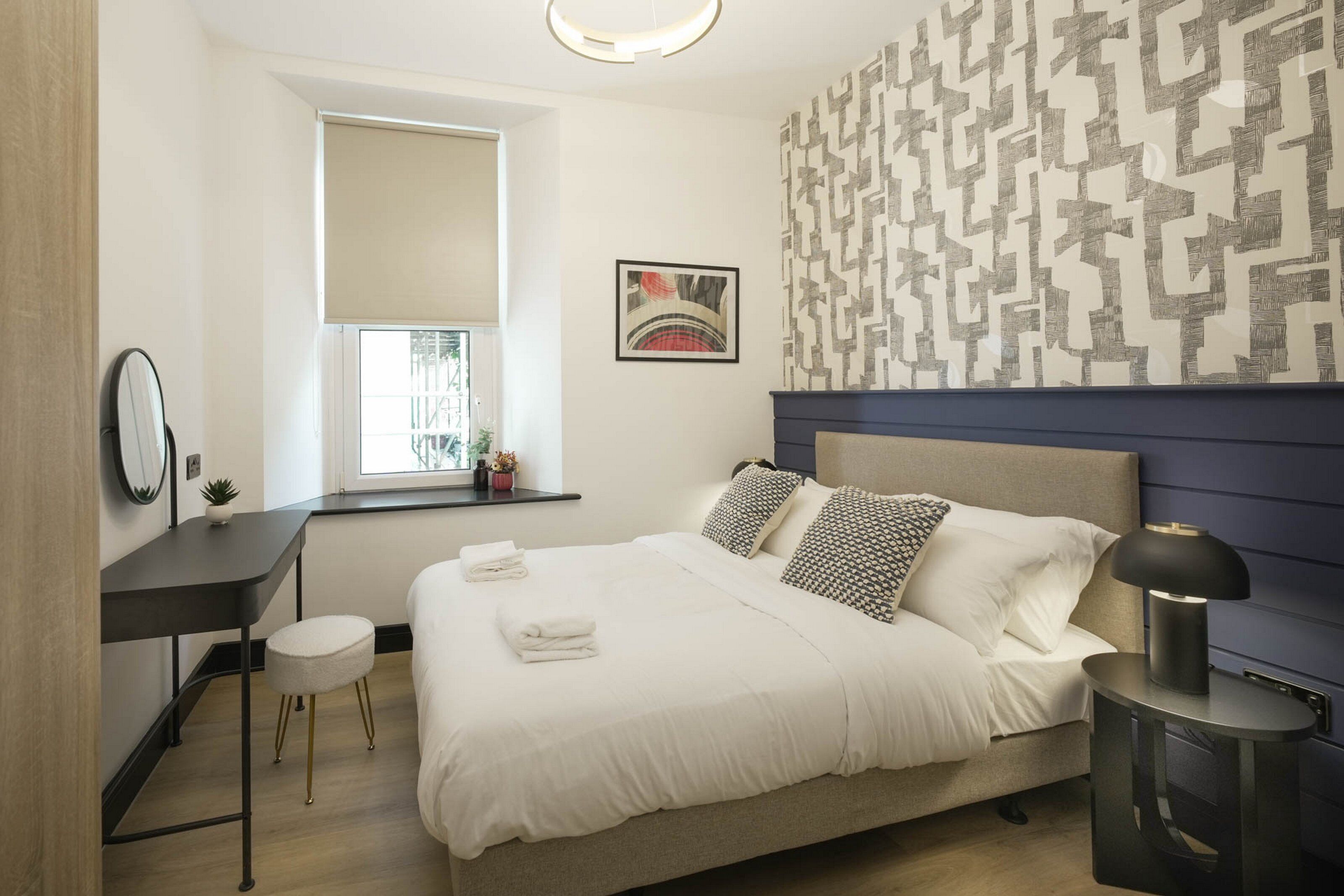 A bedroom with a bed, bedside table, lamp, window, and artwork.