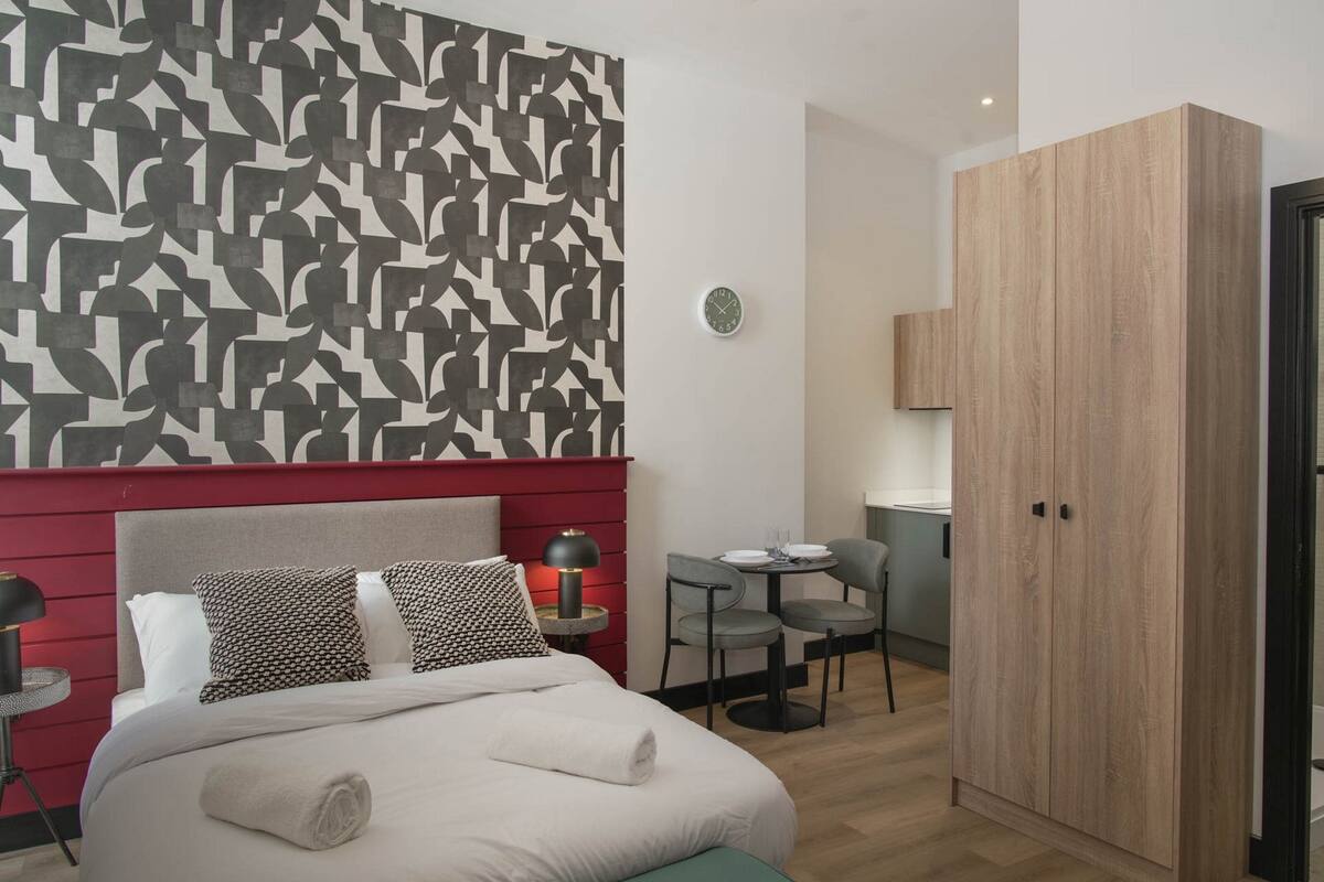 Ein modernes Hotelzimmer mit Bett, einer roten Akzentwand, Tapete mit Muster, Schreibtisch mit Stühlen und einem Kleiderschrank.