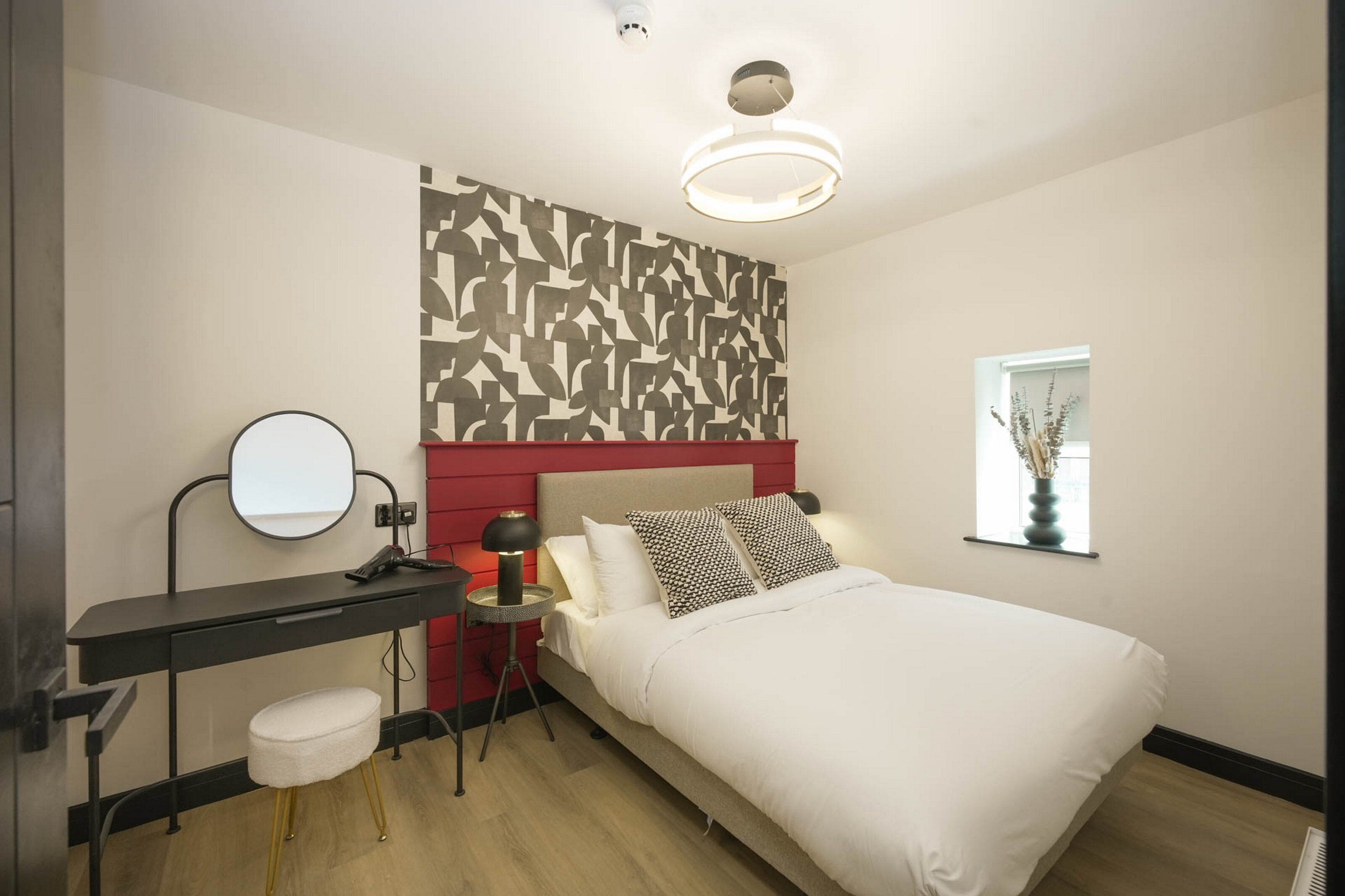 A bedroom with a bed, a desk with a mirror, a red headboard, a wall-mounted light fixture, and a window with a plant.