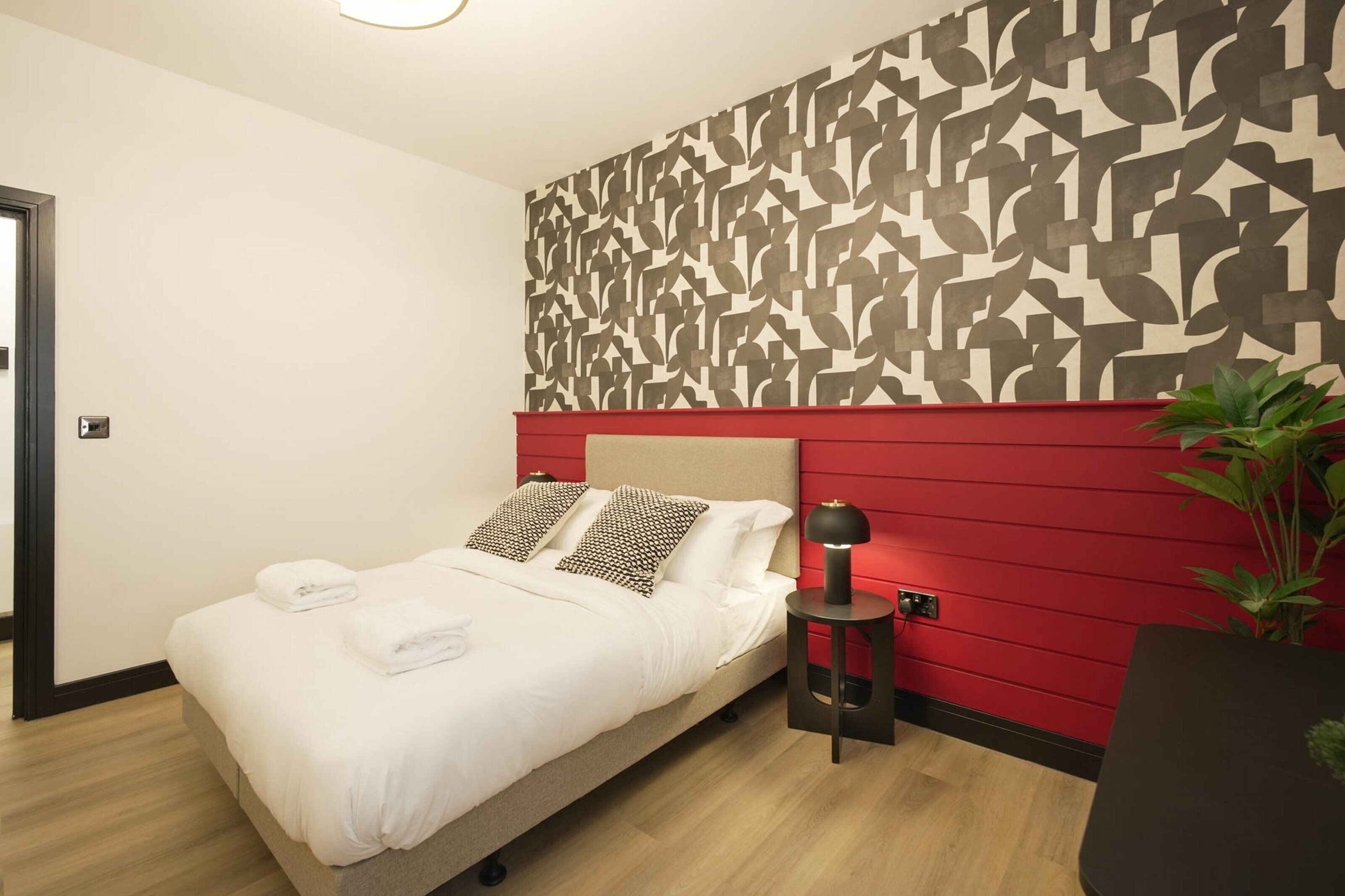 A hotel room with a bed, a red accent wall, a patterned wall, a small side table with a lamp, and a potted plant.