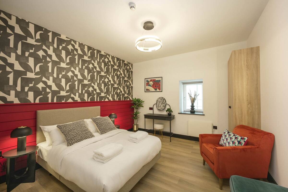 Ein modernes Hotelzimmer mit einem Bett, einem roten Kopfende, einem roten Sessel, einem kleinen Tisch und einer Wand mit geometrischem Muster.