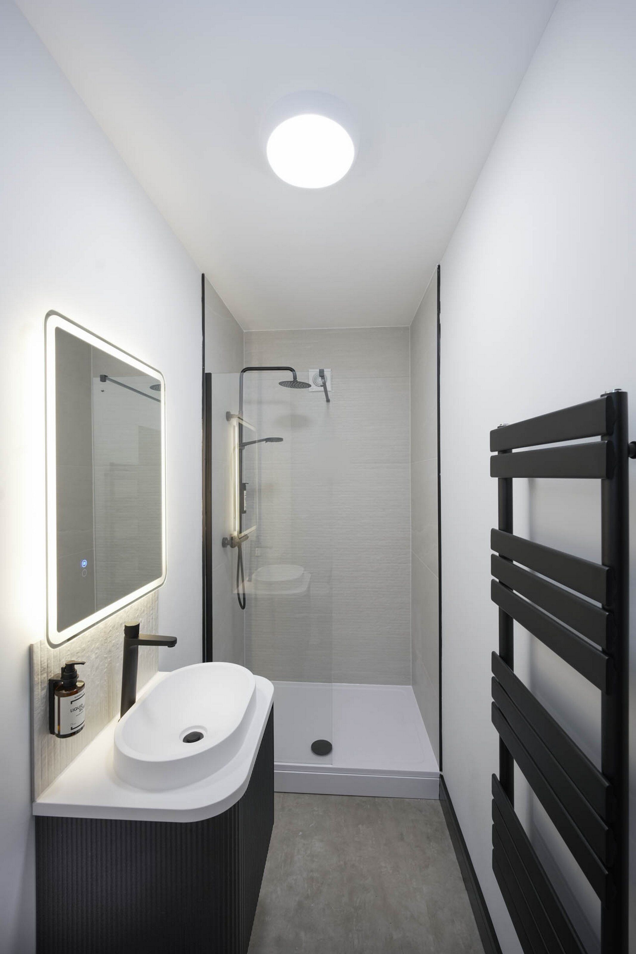 Un bagno moderno con un lavandino bianco, uno specchio, una doccia e un porta asciugamani nero.