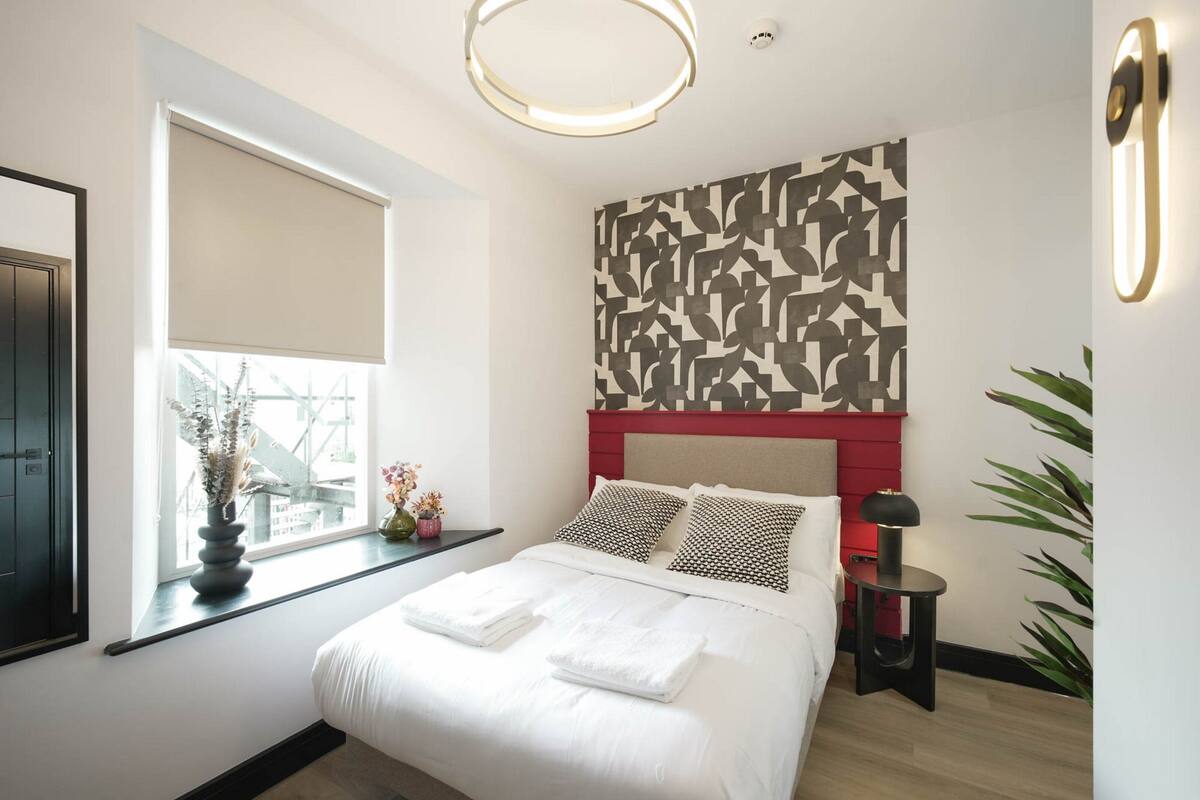 Ein modernes Hotelzimmer mit einem großen Bett, einem Fenster mit Blick, einer dekorativen Wandverkleidung und einem kleinen Tisch mit einer Lampe.