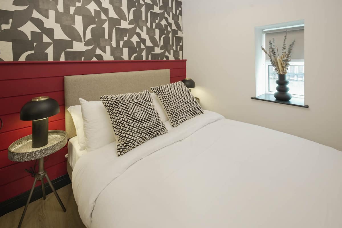 Ein Hotelzimmer mit Bett, Nachttisch mit Lampe, Fenster mit Blick und Wand mit geometrischem Muster.