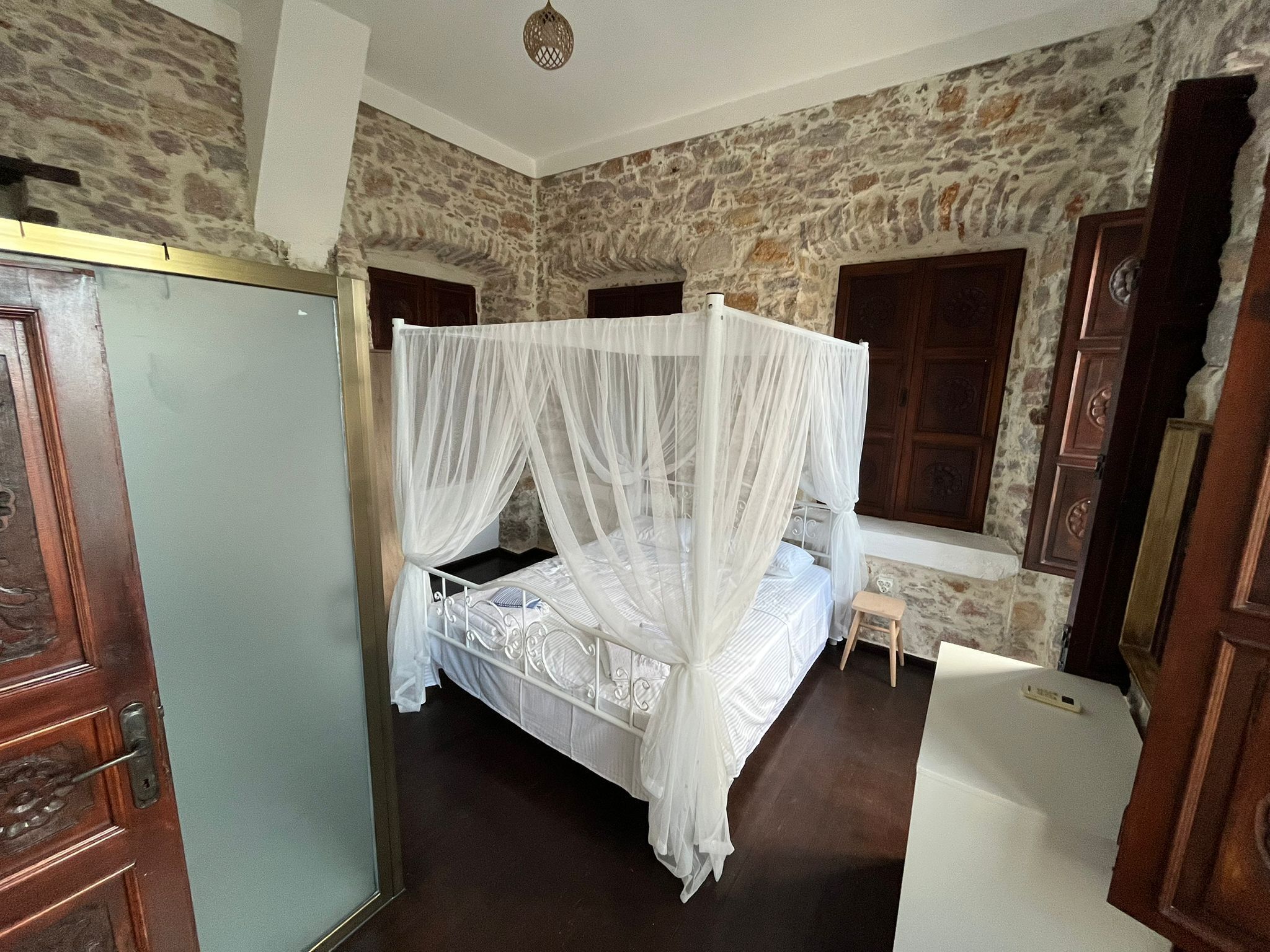 A room with a stone wall, a bed with a white canopy, a wooden door, and a small stool.
