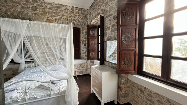 Une chambre avec un mur en pierre, un lit à baldaquin, une commode et une fenêtre à carreaux.