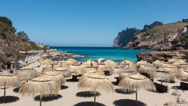 Ein Strand mit Reetsonnenstühlen, blauem Meer, steilen Klippen und Gebäuden am Hang.