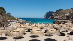 A beach with thatched umbrellas, a clear blue sea, rocky cliffs, and buildings on the hillside. - Beautiful house with WIFI, air conditioning and pool in exclusive urbanization (Cala San Vicente, Pollensa)