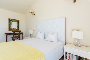 A hotel room with a large bed, a bedside table with a lamp, a small desk with a chair, and a bookshelf with books. - Jardines de Calahonda IX | Sea View & Pool (Mijas Costa)