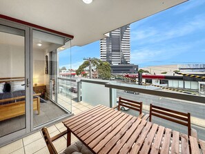 A balcony with a wooden table and chairs, a bed with a glass door, and a view of a cityscape with buildings and a road. - Heart of Geelong Near Waterfront and Train (Geelong)