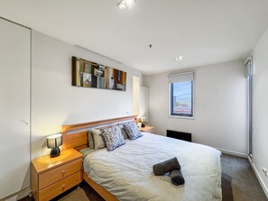 Un dormitorio con cama, mesitas de noche, lámpara, un cuadro y una ventana.